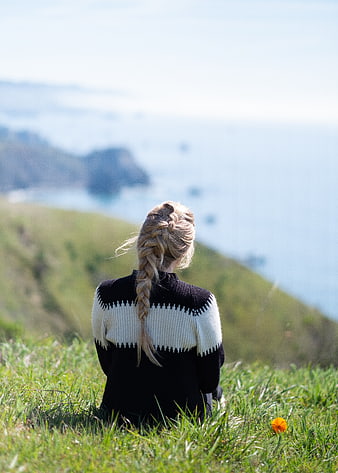 Blonde Woman Sitting on Grassy Hill Overlooking Ocean Coast