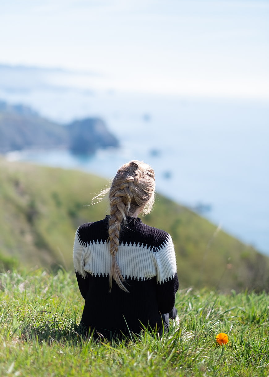 Blonde Woman Sitting on Grassy Hill Overlooking Ocean Coast