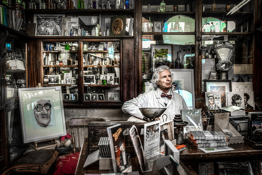 Man in White Coat Sitting in Cluttered Antique Shop