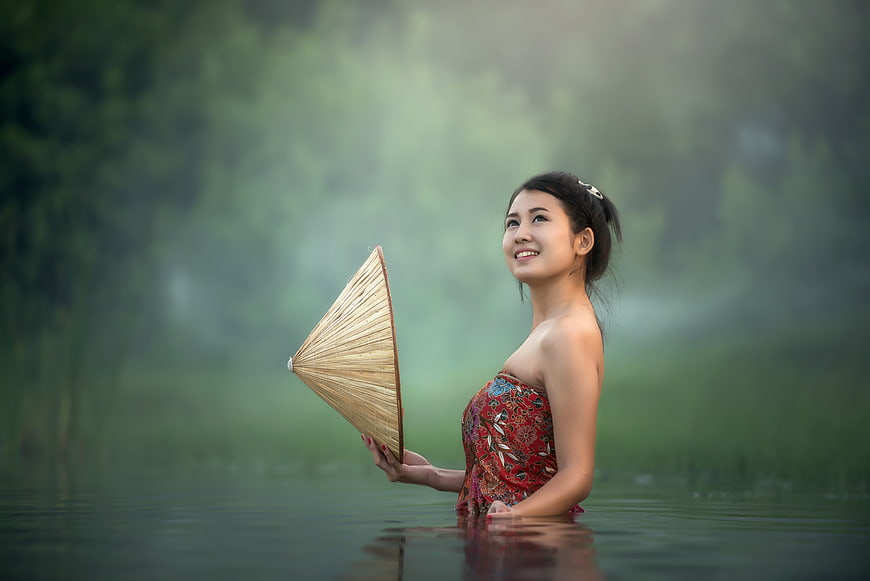 Asian woman in red dress holding conical hat in misty lake