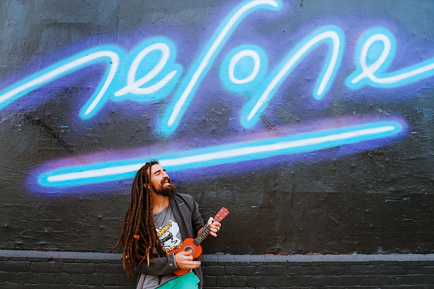Man with dreadlocks playing ukulele against neon graffiti wall