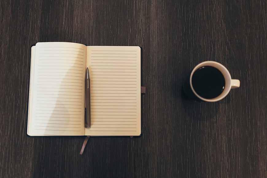 Flat lay of open notebook, pen, and coffee cup on dark wood