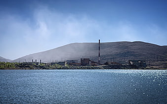 Industrial plant by lake with smokestack and mountain backdrop