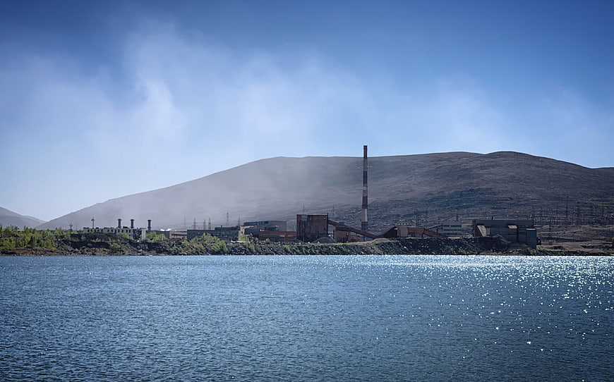 Industrial plant by lake with smokestack and mountain backdrop