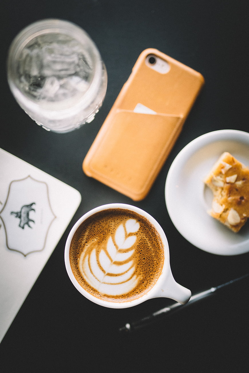 Flat lay of latte art, water, and almond bar on dark table