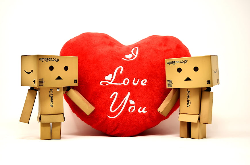 Two cardboard robots holding a red heart pillow with Love You text