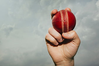 Hand Holding Red Cricket Ball Against Cloudy Sky