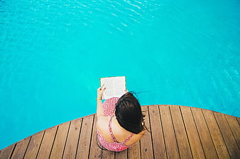 Woman reading book by turquoise pool on wooden deck