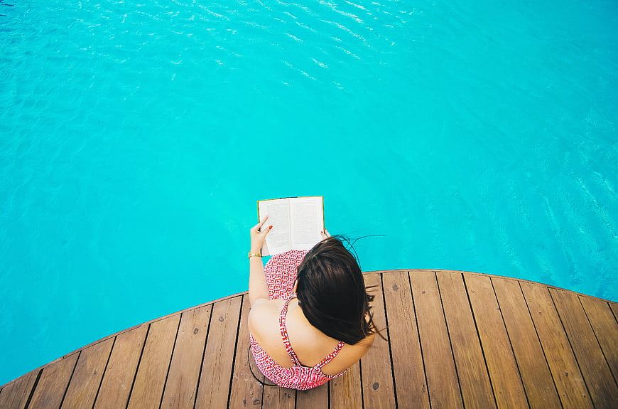 Woman reading book by turquoise pool on wooden deck
