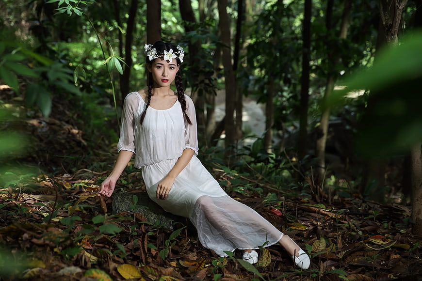Young woman in white dress sitting in forest with flower crown