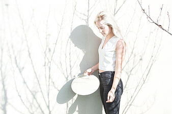 Blonde Woman in White Top Holding Hat Against White Wall