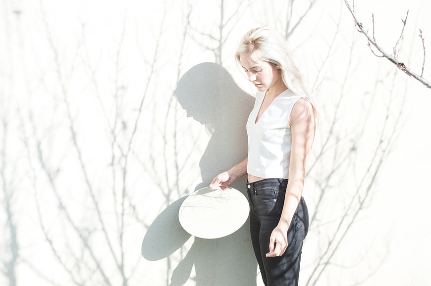 Blonde Woman in White Top Holding Hat Against White Wall