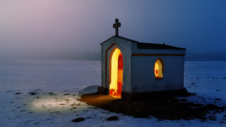 Small illuminated chapel in snowy winter landscape at dusk