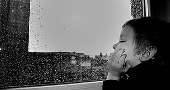 Sad child looking out rainy window in black and white