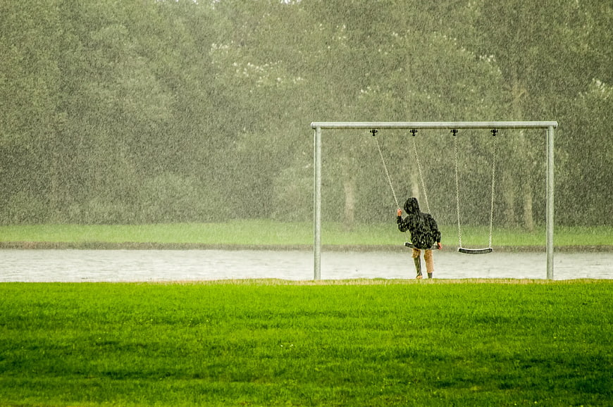 Child in black hoodie swinging in heavy rain near lake