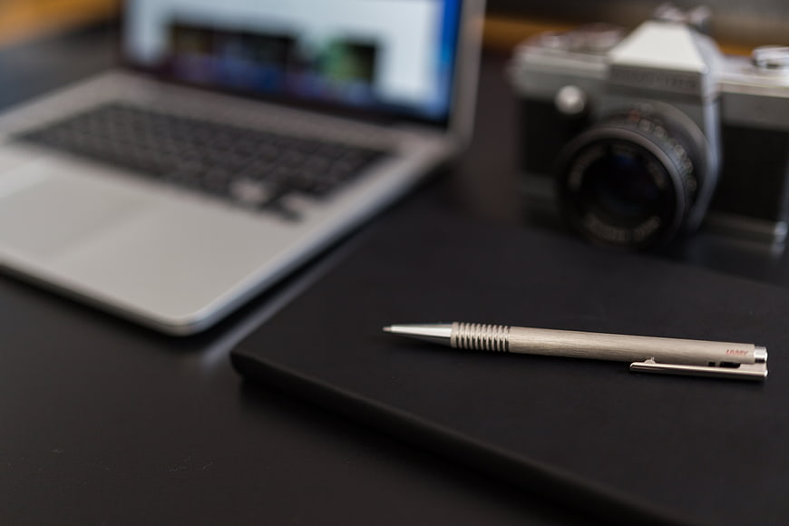 Silver mechanical pen on black notebook with laptop and camera