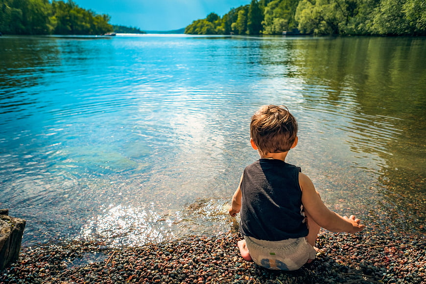 Toddler Boy Sitting by Lake Shore in Summer