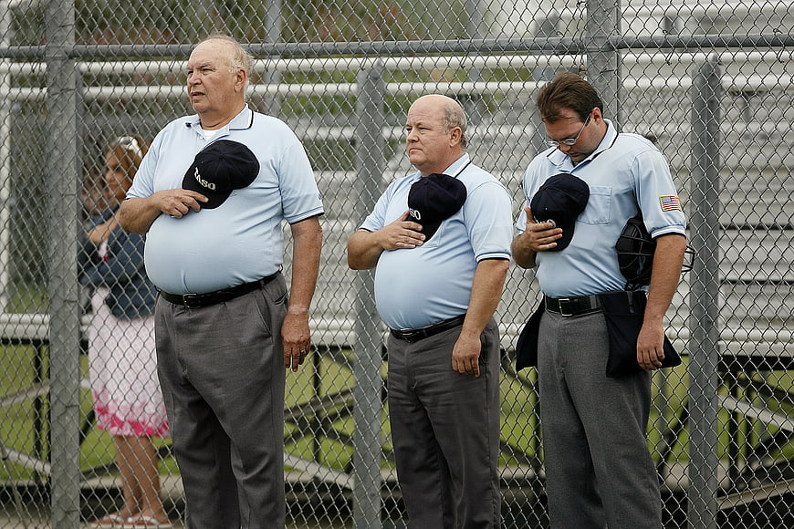 Three Umpires Standing At Chain-Link Fence During Softball Game