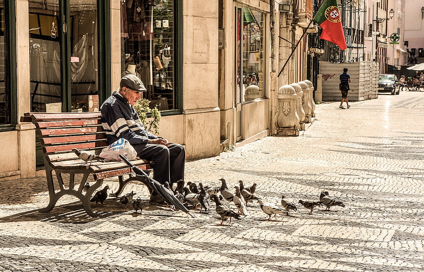 Elderly man sitting on a bench feeding pigeons on a Portuguese street