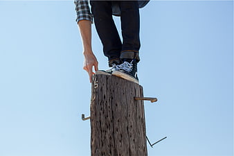 Man standing on wooden utility pole against blue sky