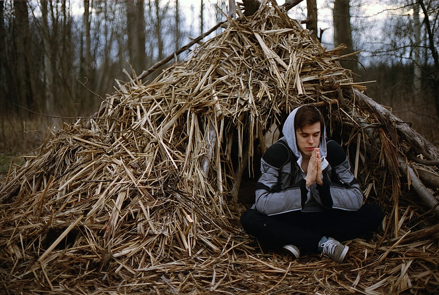 Young man meditating in front of rustic straw hut in forest