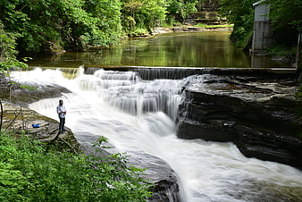 Man fishing on rocky ledge beside cascading river waterfall in lush green forest