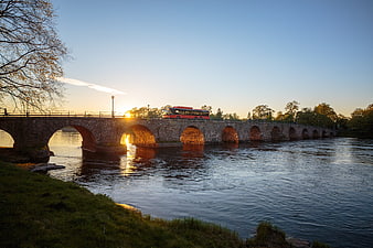 Red bus crossing stone arch bridge over river at sunset in Sweden