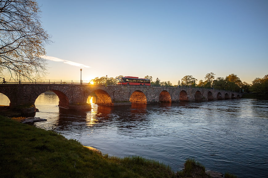 Red bus crossing stone arch bridge over river at sunset in Sweden