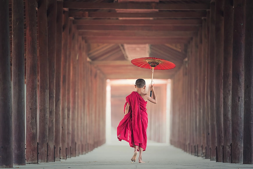 Young Buddhist Monk Walking with Red Umbrella in Wooden Corridor