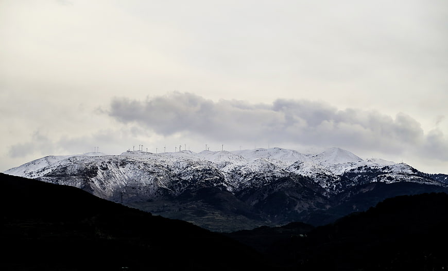 Snow-Capped Mountain Range with Wind Turbines Under Cloudy Sky