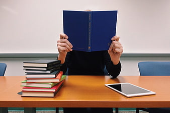 Student Reading Book at Desk with Tablet and Stack of Textbooks