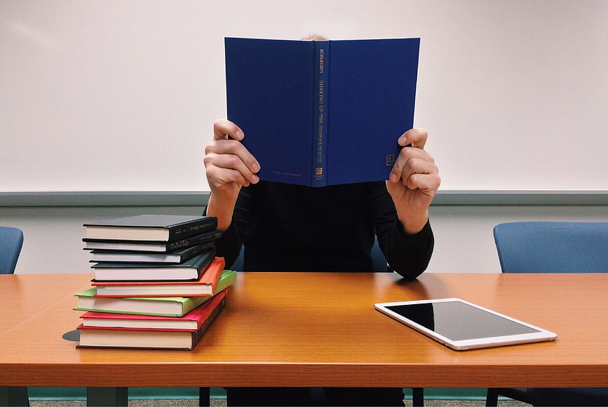 Student Reading Book at Desk with Tablet and Stack of Textbooks