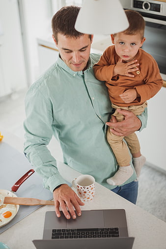 Father working on laptop while holding toddler in kitchen