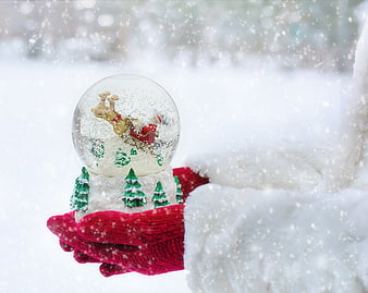Hand in red glove holding Christmas snow globe in winter snow