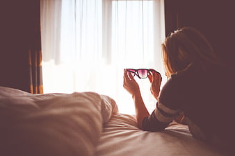 Woman holding sunglasses in bed near bright window