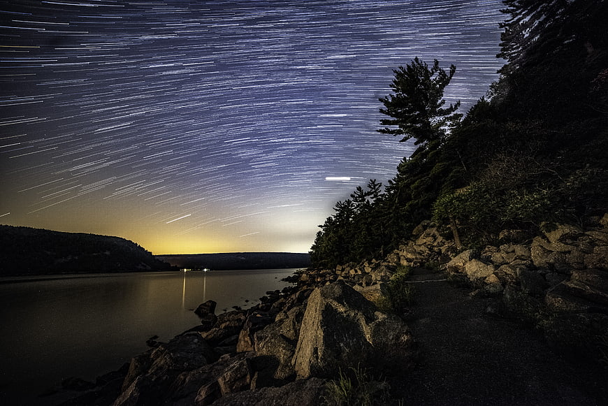 Star trails over a calm lake at night with rocky shoreline