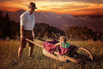 Senior man pulling wooden cart with young girl in mountain meadow at sunset