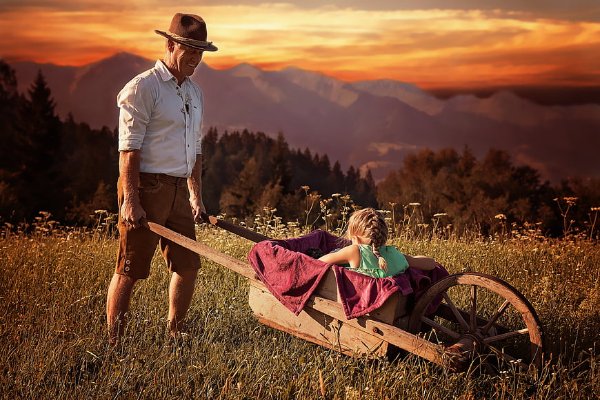 Senior man pulling wooden cart with young girl in mountain meadow at sunset