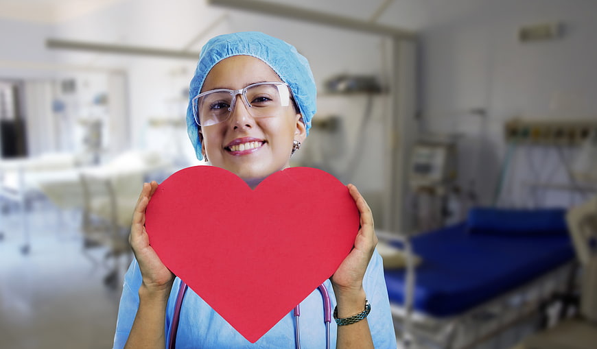 Smiling female nurse holding red heart in hospital ward