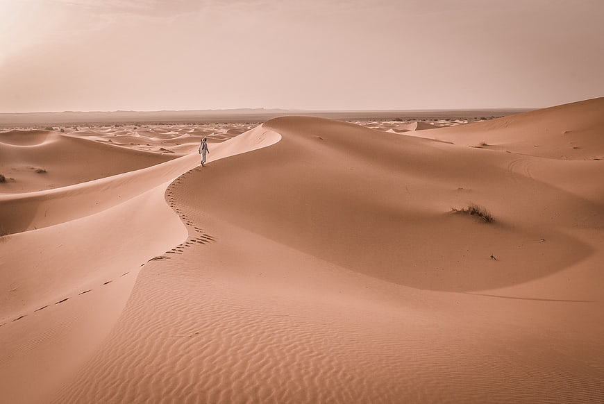 Lone figure walking on vast desert sand dunes with footprints