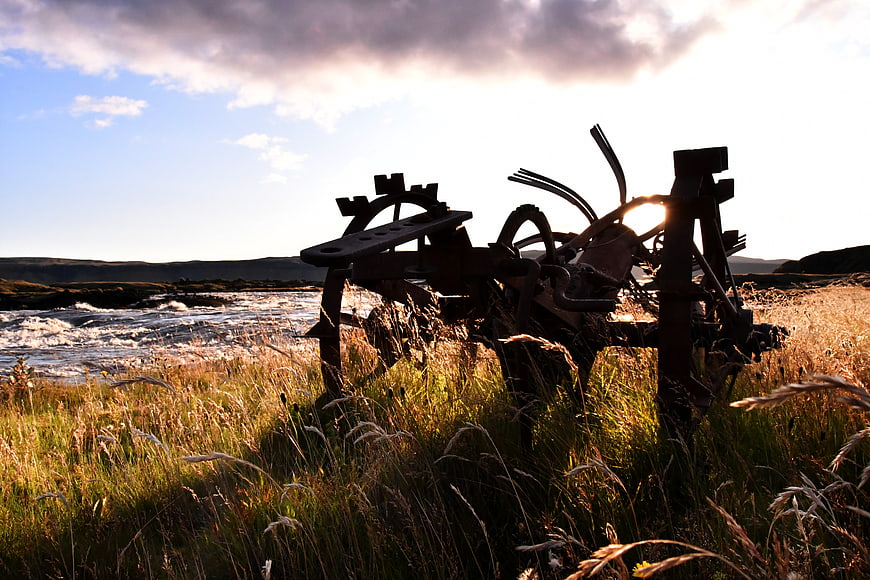Rusty Farm Plow Silhouette in Grassy Field by River