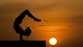 Woman performing yoga handstand silhouette against orange sunset sky