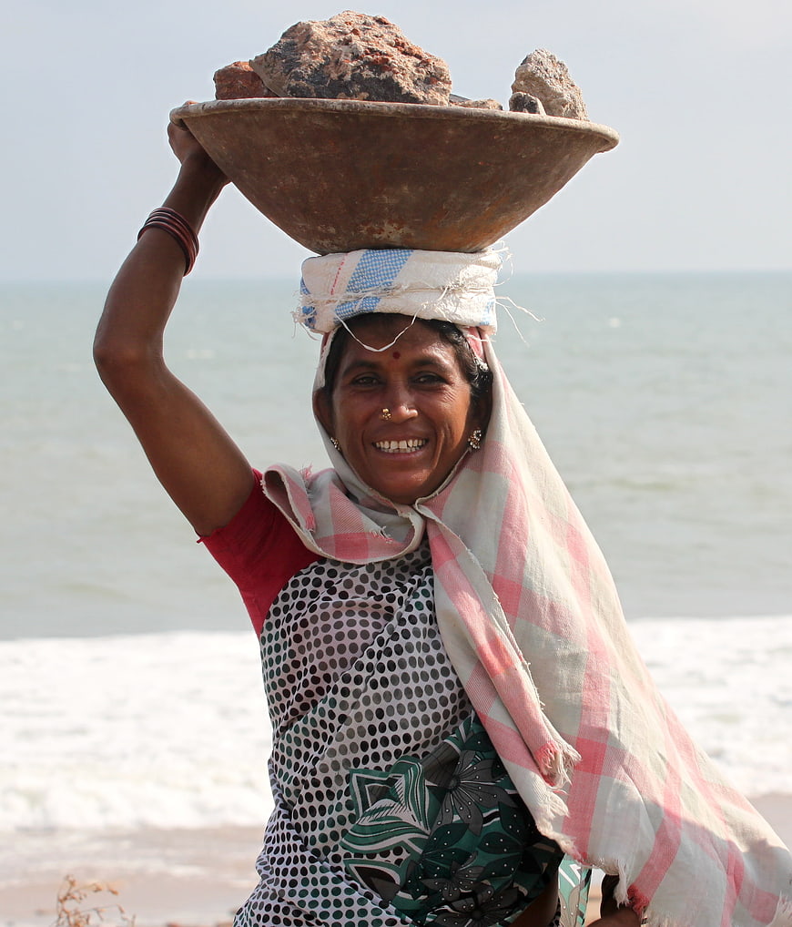 Smiling Indian woman carrying heavy rocks on her head at the beach