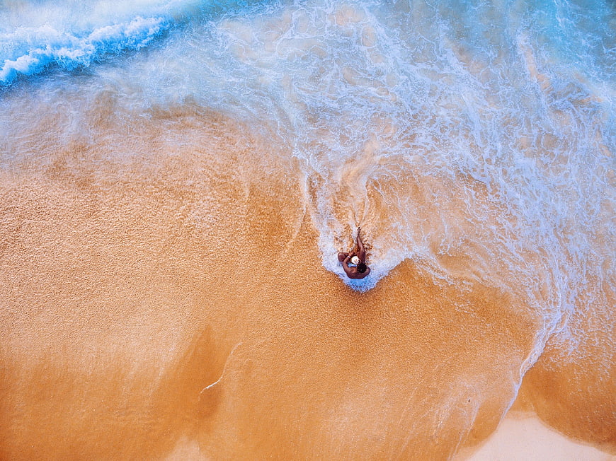 Aerial view of man walking in ocean waves on sandy beach