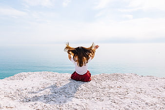 Woman sitting on cliff overlooking ocean with arms raised