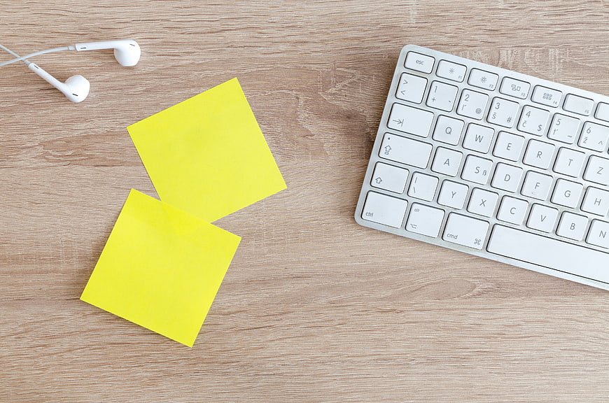 Flat lay of yellow sticky notes, white keyboard, and earbuds on wooden desk