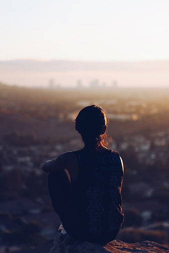 Woman sitting on cliff overlooking city at sunset