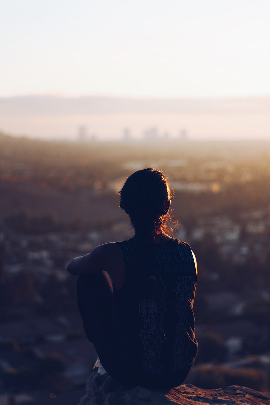 Woman sitting on cliff overlooking city at sunset