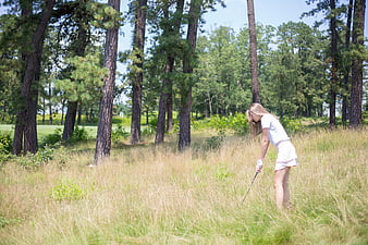 Woman golfer playing in tall grass near pine trees