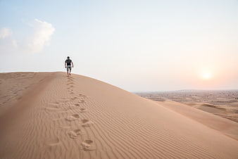 Man Walking Up Sand Dunes In Desert At Sunset